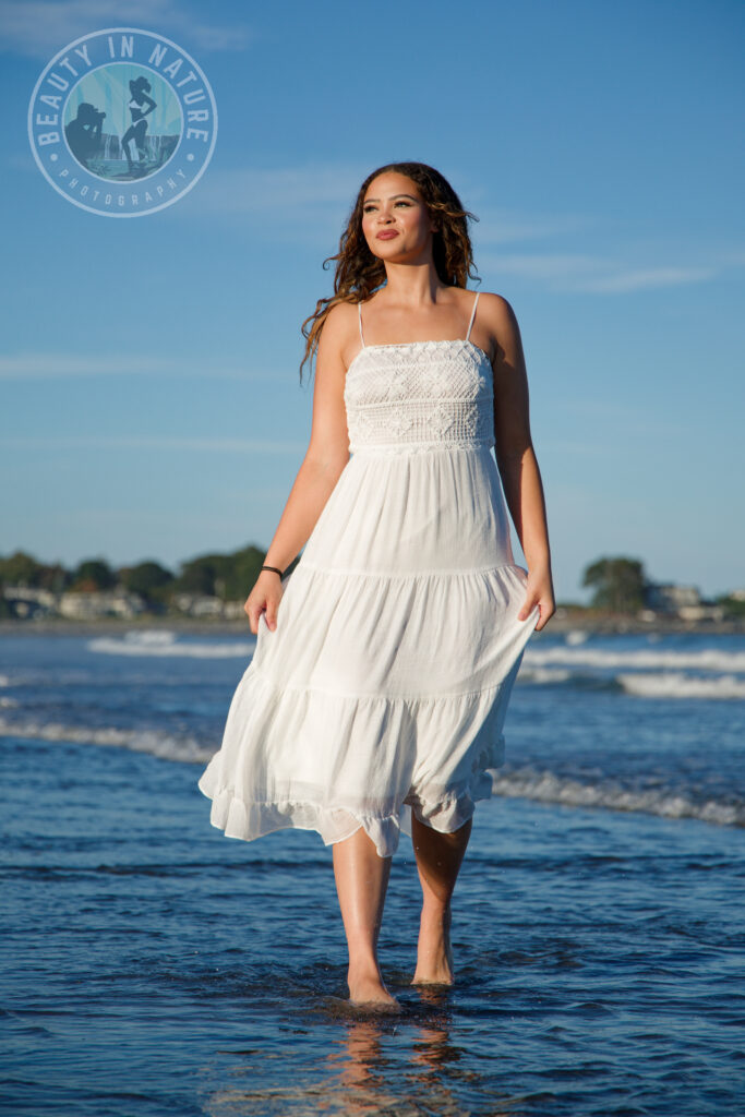 Beautiful high school senior girl walking along the beach in white, flowy dress