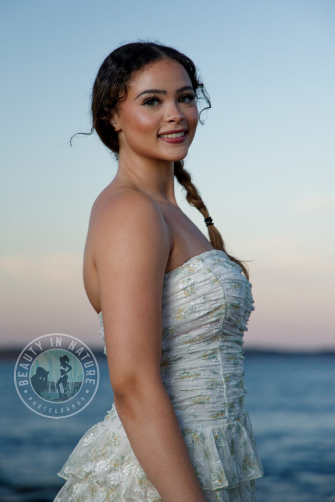 Beautiful high school senior girl in party dress posing at shoreline at sunset